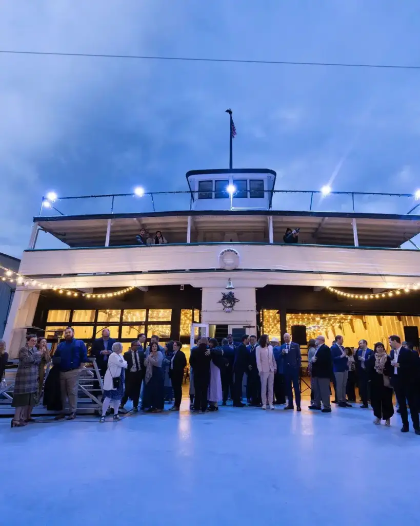 Guests on the deck of the MV Skansonia at dusk, with the lit ferry cabin and string lights behind them on Lake Union