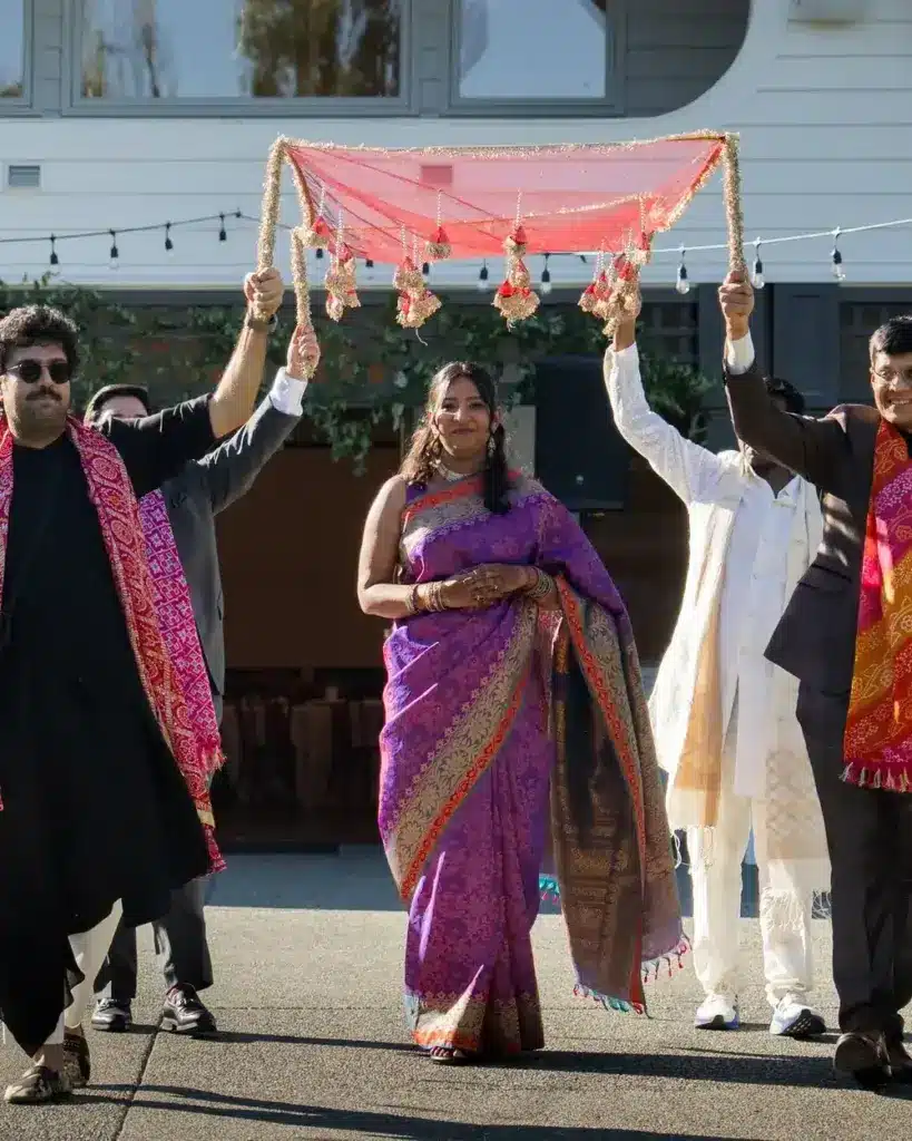 A bride in a purple silk sari walking beneath a red phulkari canopy held by her family outside the winery hall at Columbia Collective in Woodinville