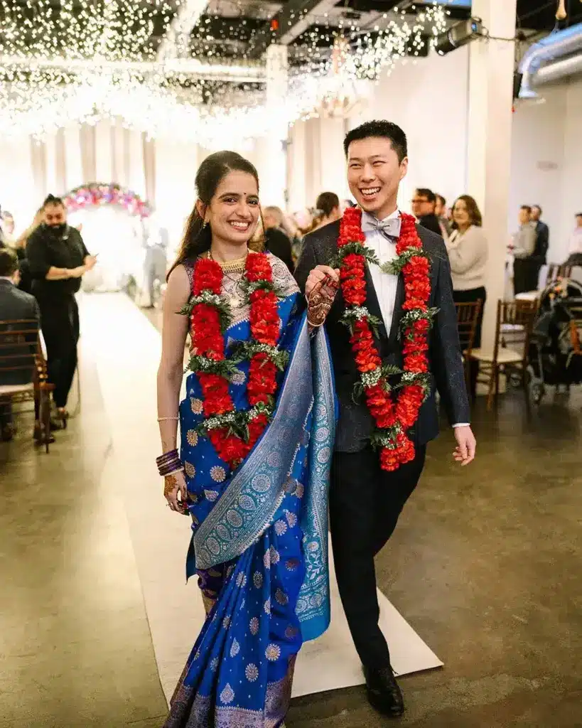 Newlyweds in red flower garlands walking up the aisle beneath a ceiling of lights at Hideaway Cellars