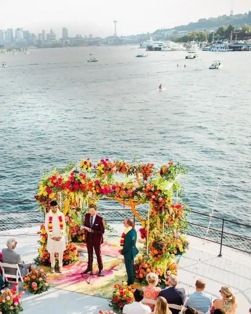 A wedding ceremony beneath a marigold and rose mandap on the bow of the MV Skansonia, with Lake Union and the Seattle skyline behind