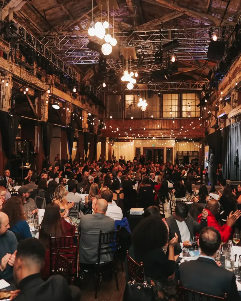 A packed gala dinner in the SoDo Park hall, with guests applauding beneath the timber trusses and a presentation on screen