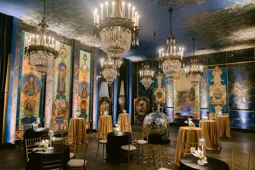The muraled ballroom at The Ruins set for an evening party, with gold-draped cocktail tables beneath crystal chandeliers