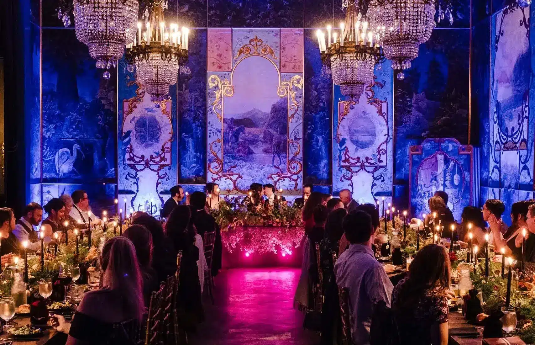 Long candlelit dinner tables beneath crystal chandeliers in the muraled ballroom at The Ruins, a Seattle event venue