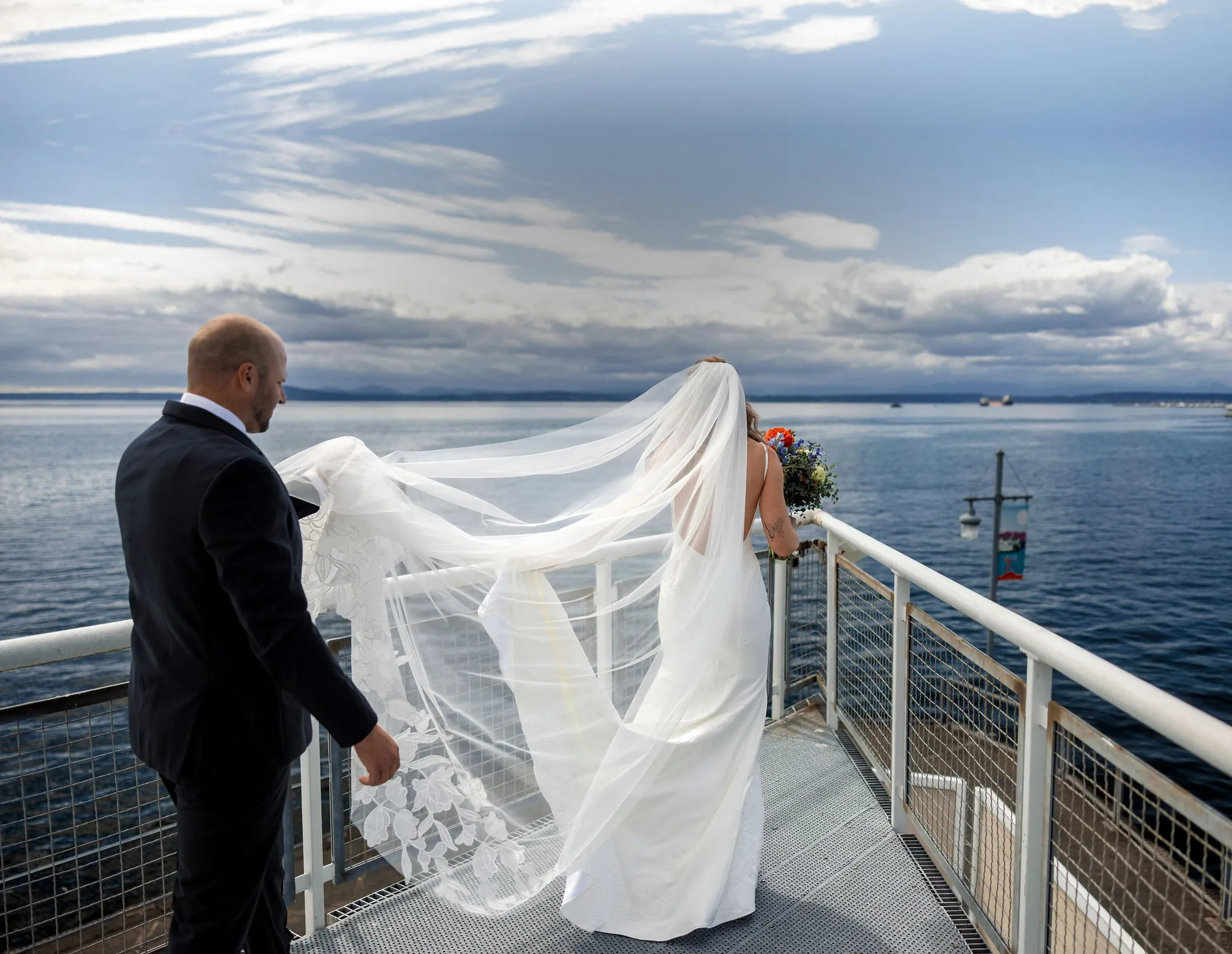 Bride and groom on the waterfront deck at The Point at Pier 70, her veil lifting over Elliott Bay, a Seattle wedding venue